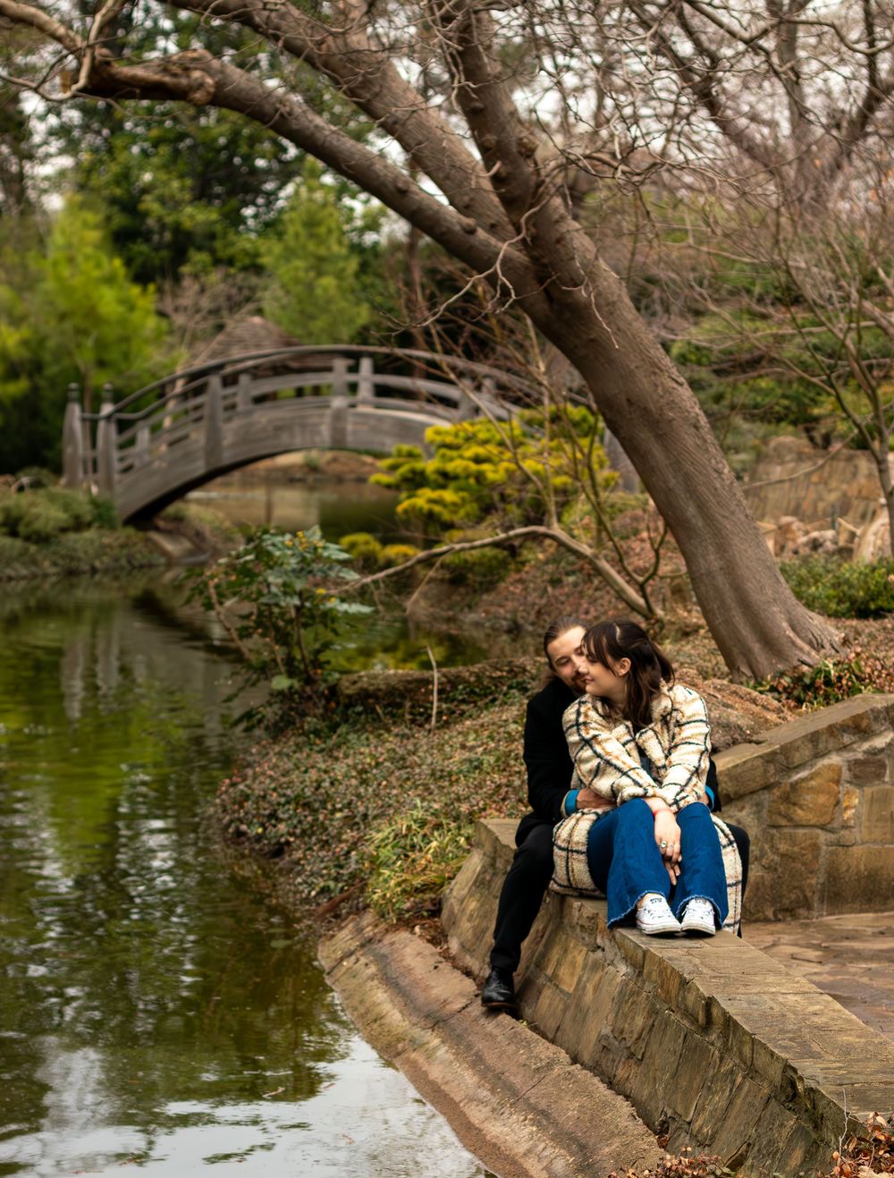 Couple sharing a romantic moment by a serene pond in a Japanese garden with a wooden bridge backdrop.