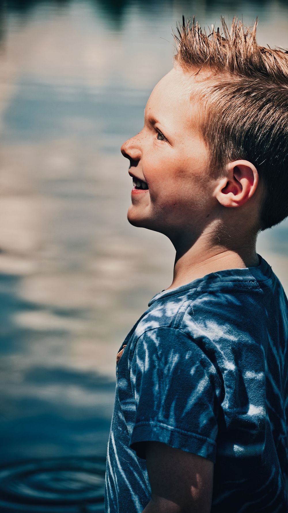 Side profile of smiling boy with spiked hair in blue tie-dye shirt near sparkling water.