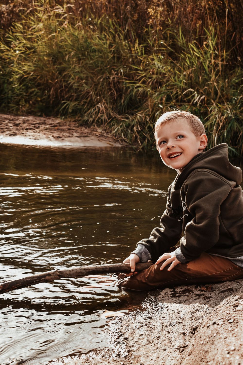 Smiling young boy in brown hoodie sits by shallow creek shore holding a stick outdoors.