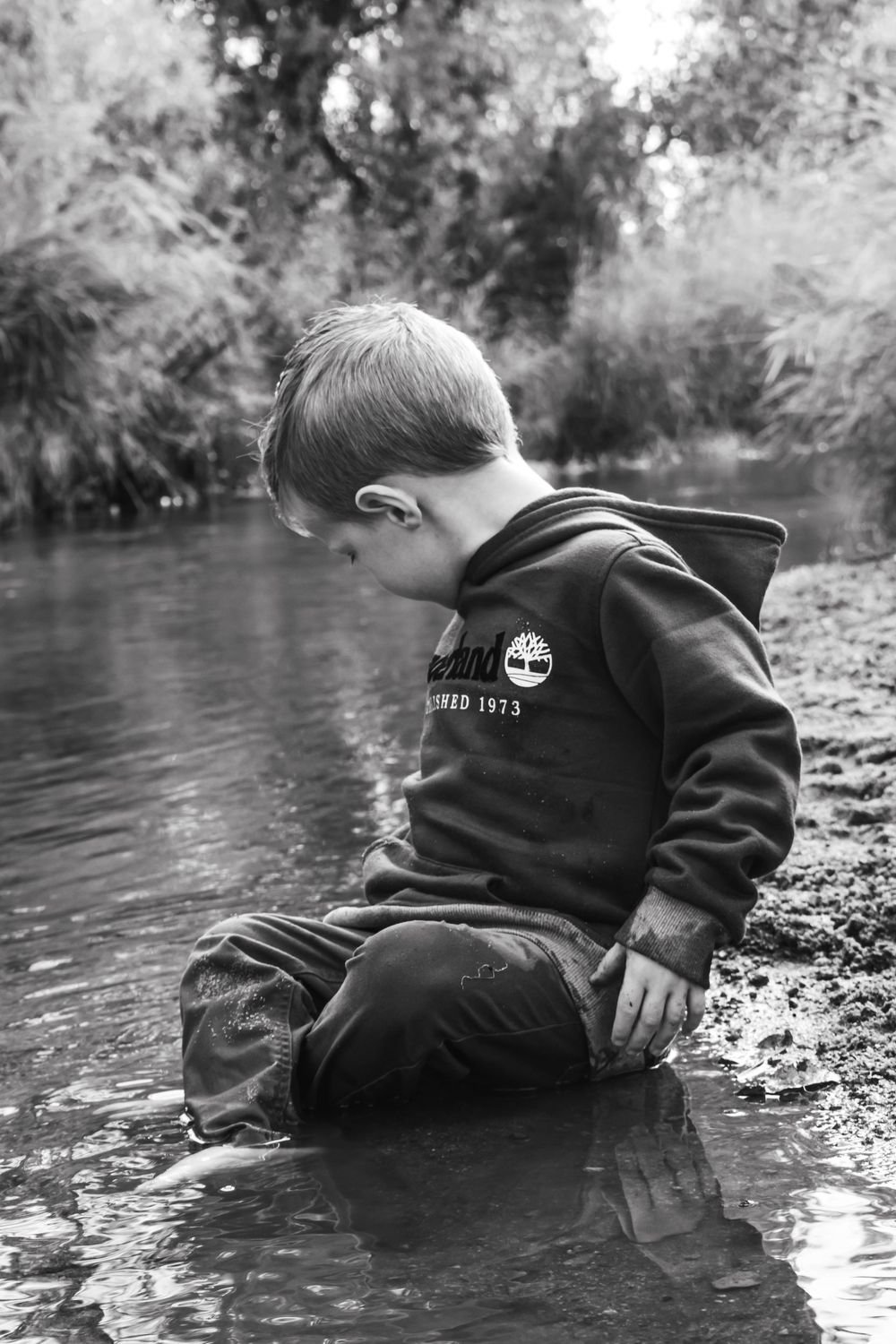 Black and white photo of young boy in Timberland hoodie sitting in shallow creek water.