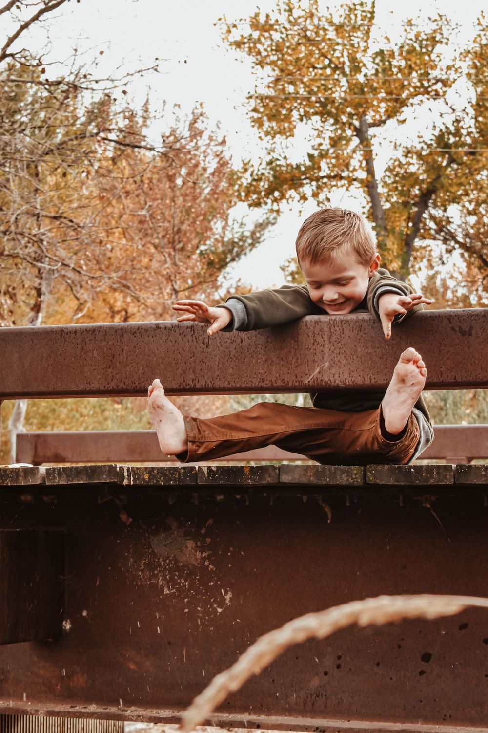Barefoot smiling boy hangs through rusty metal fence rails on bridge surrounded by autumn trees.