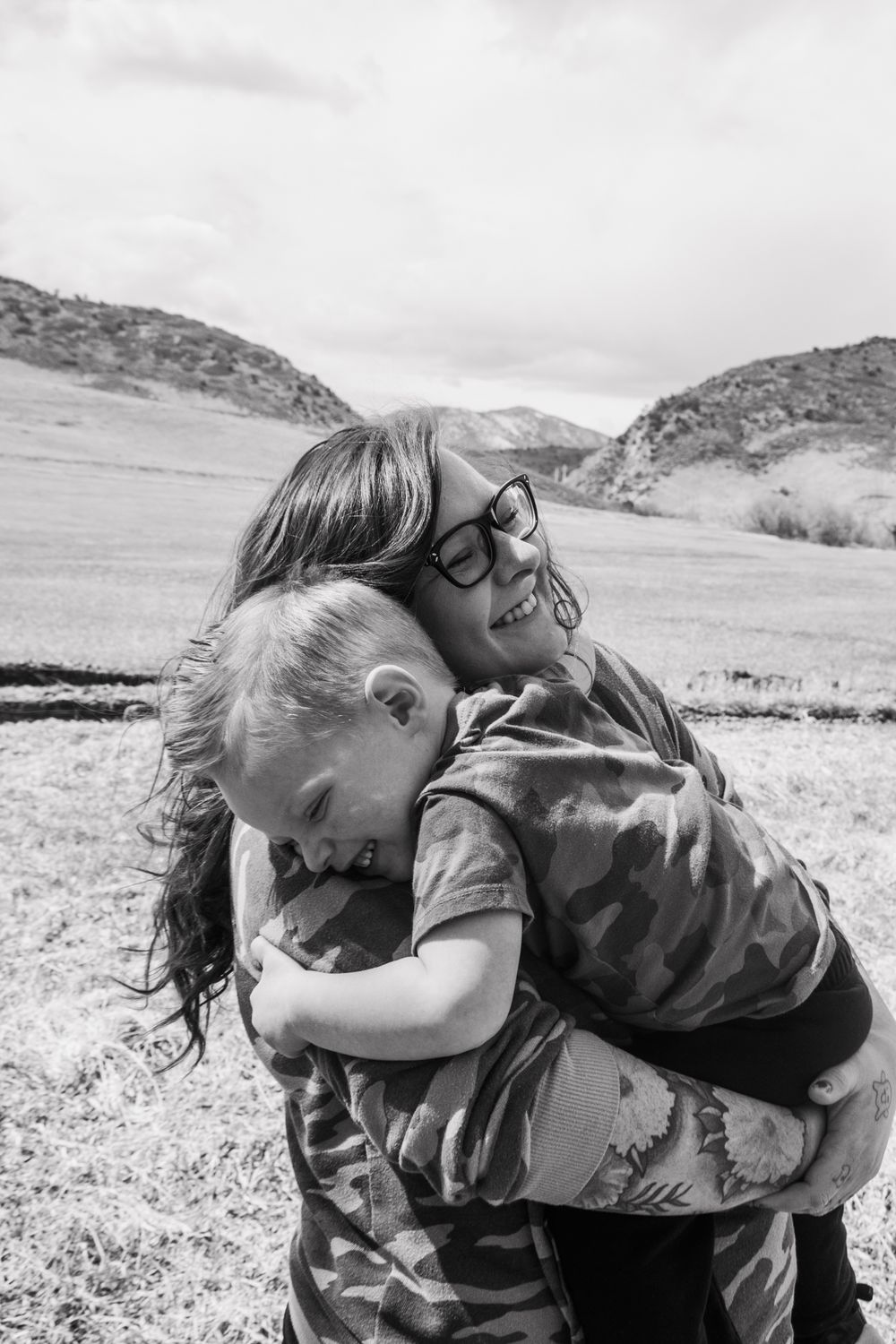 Two children hugging and smiling in a field with hills in black and white photo.