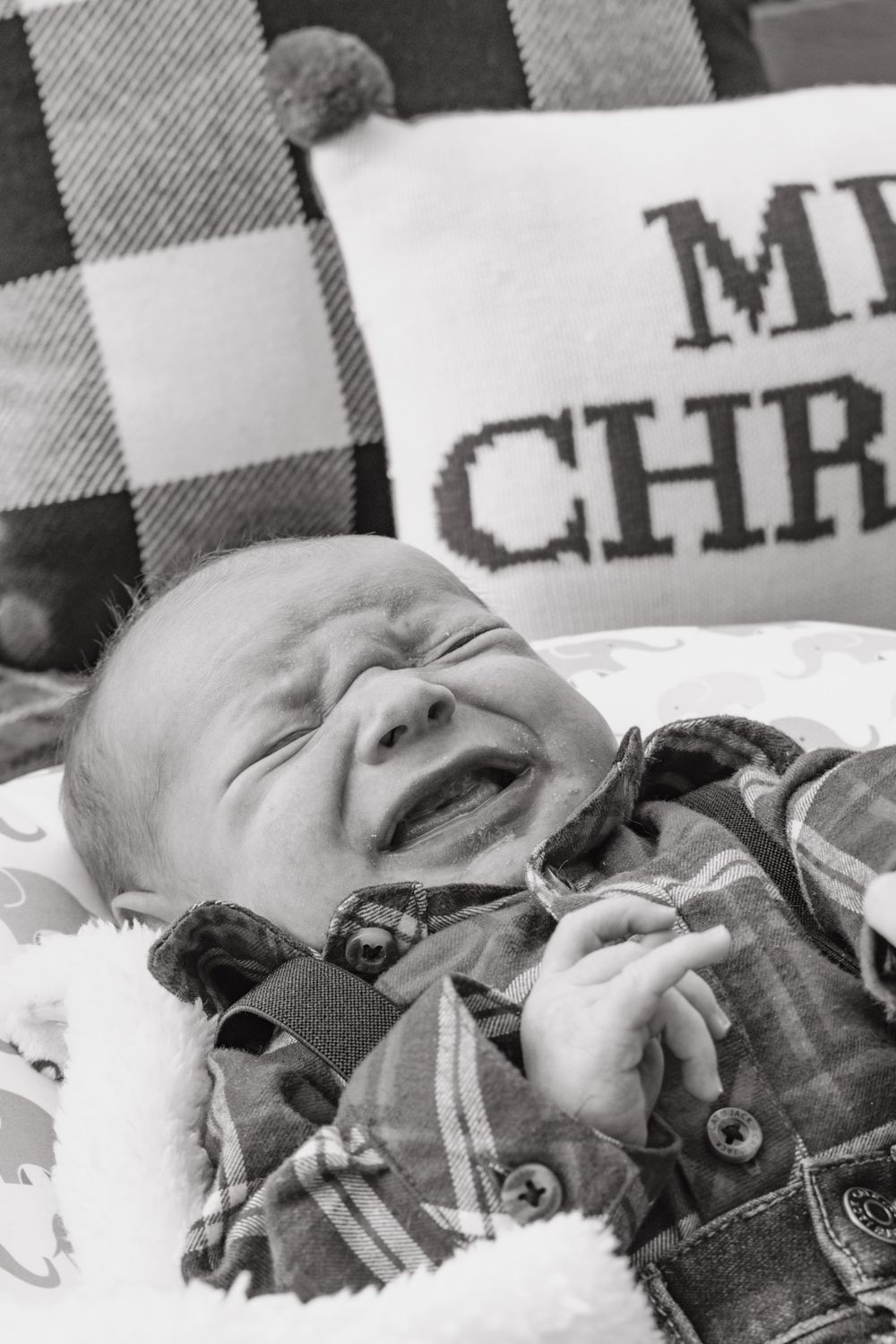 Crying newborn baby in plaid shirt with Christmas pillow in black and white photo.