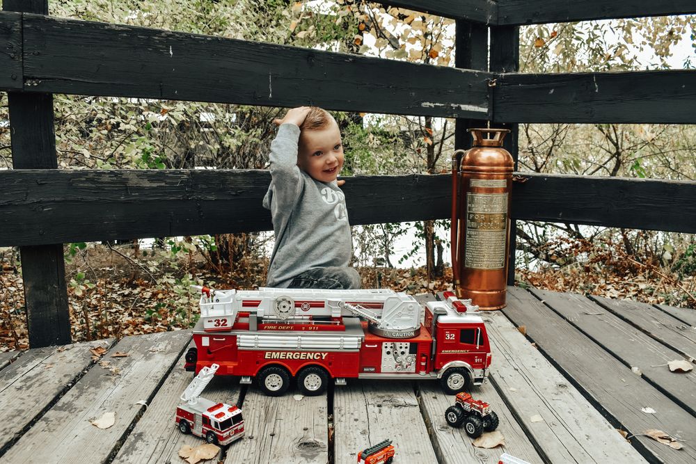 Happy toddler plays with toy fire trucks and copper fire extinguisher on wooden outdoor deck.