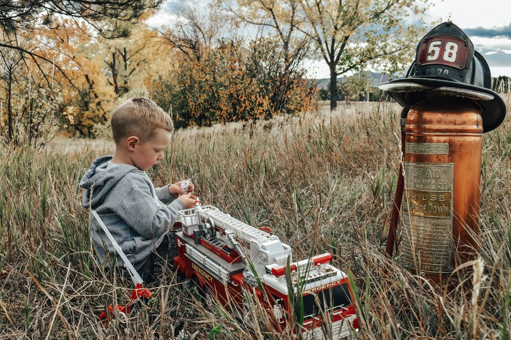 Young boy sits in tall autumn grass playing with toy fire truck next to vintage fire extinguisher.