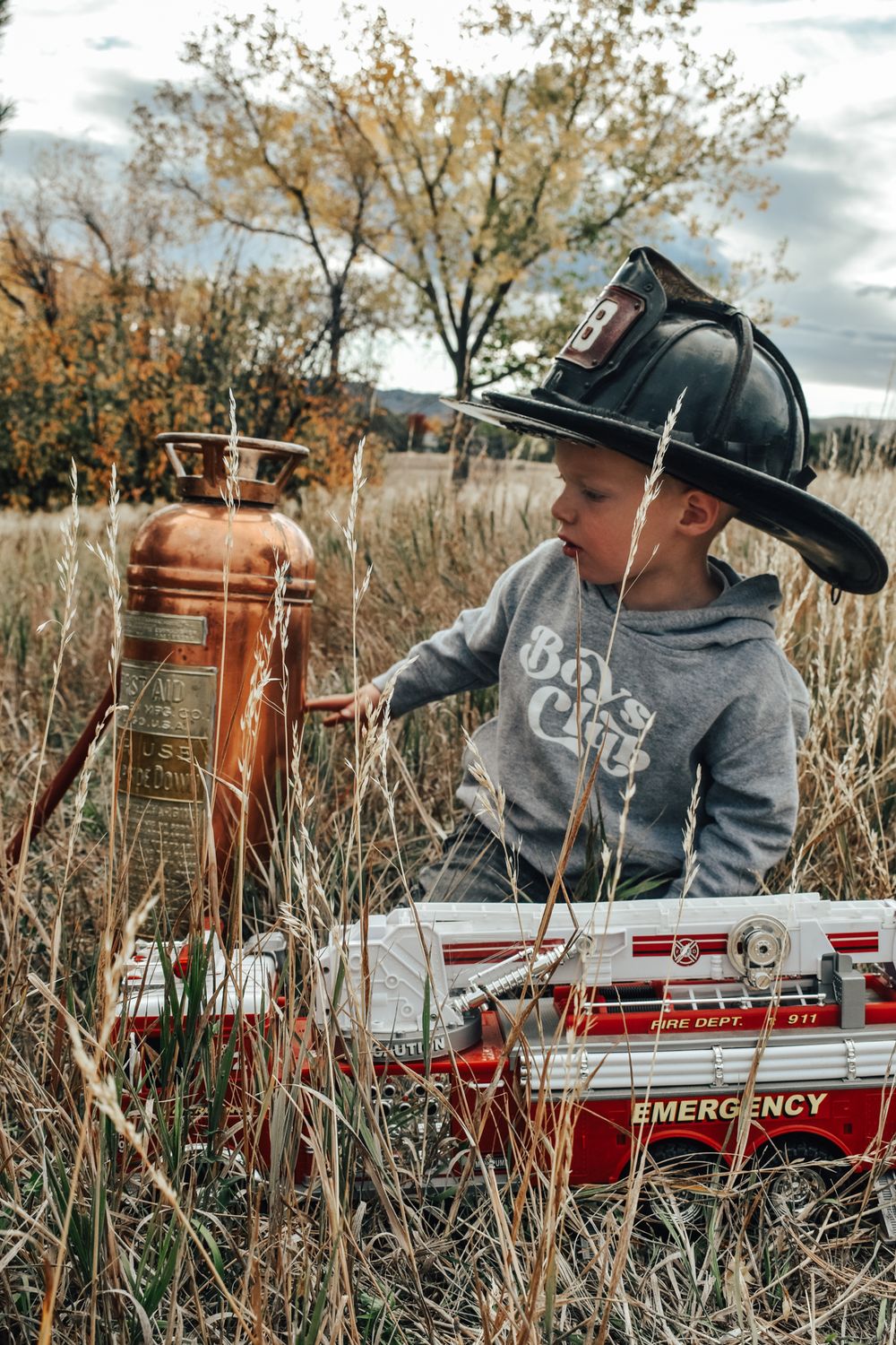 Boy wearing firefighter helmet examines vintage fire extinguisher beside toy fire truck in tall grass.