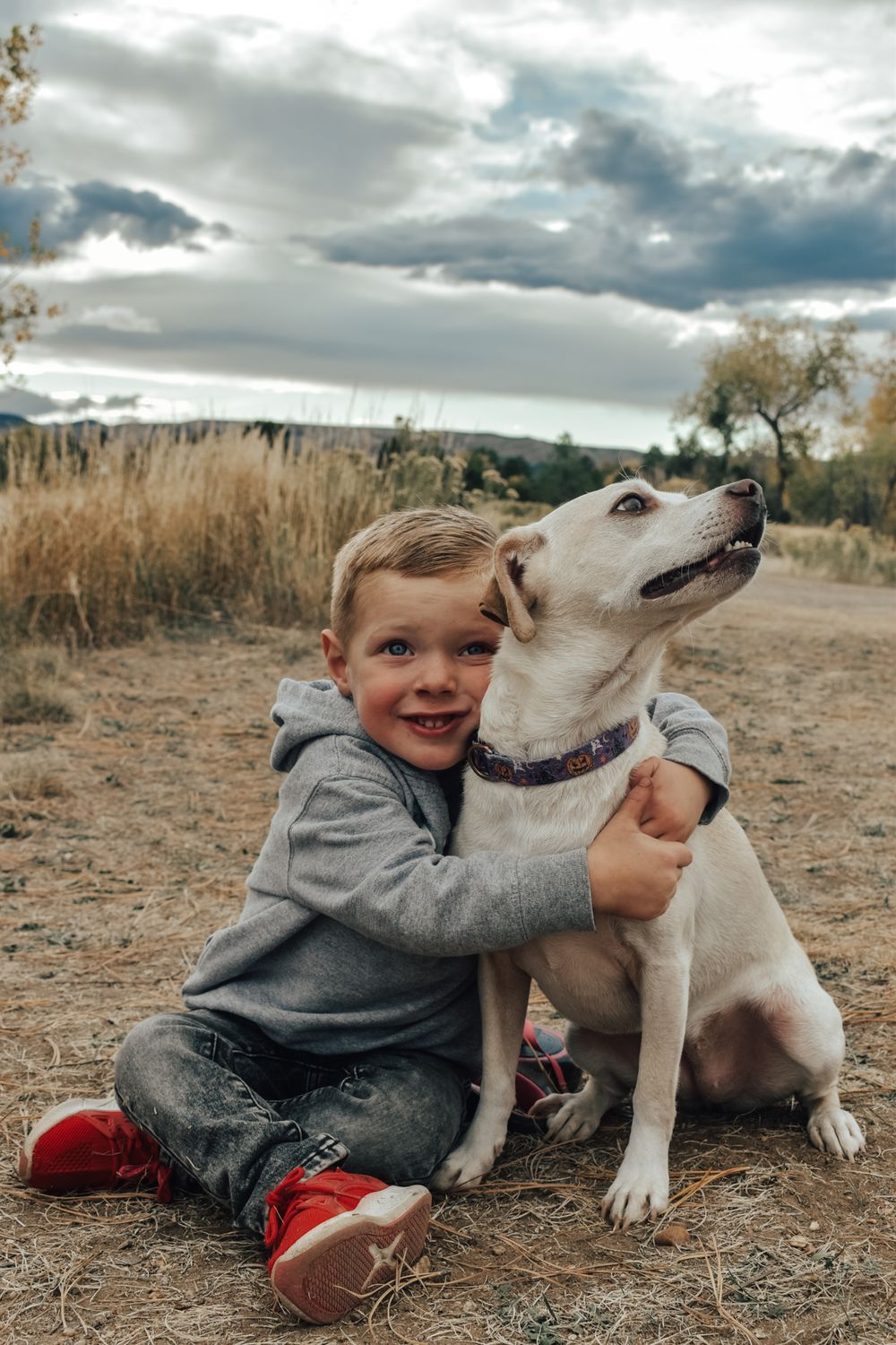 Smiling young boy hugging a white dog while sitting on dry grass outdoors.