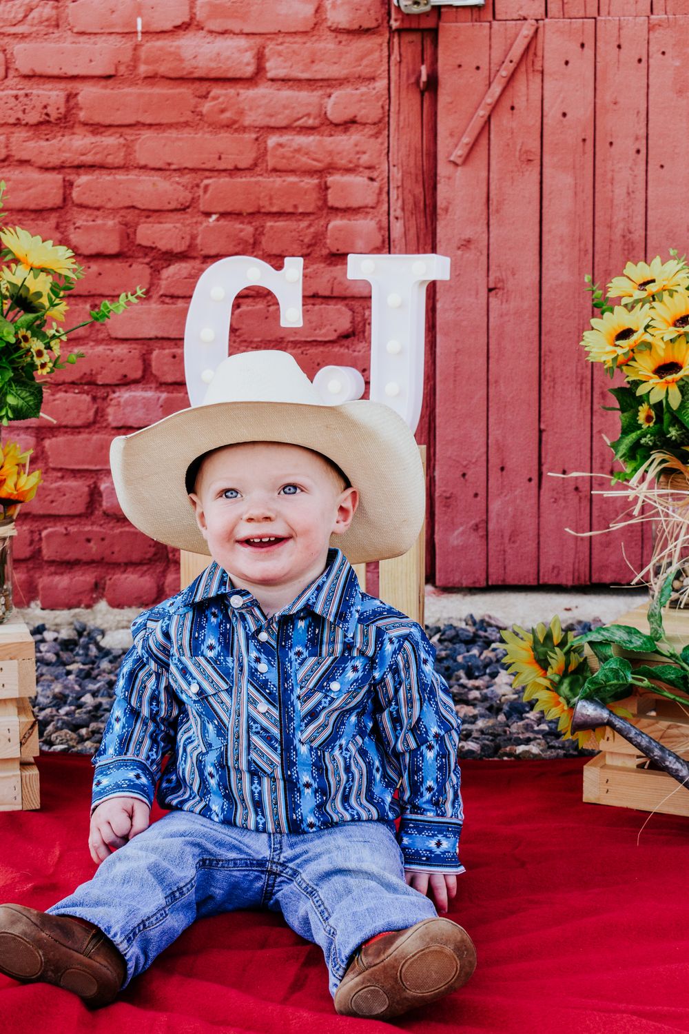 Happy baby boy in cowboy hat and blue western shirt sits before red brick wall with CJ letters.