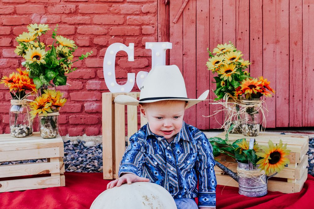 Toddler boy wearing white cowboy hat sitting near rustic red barn with sunflowers and wooden crates at birthday party.