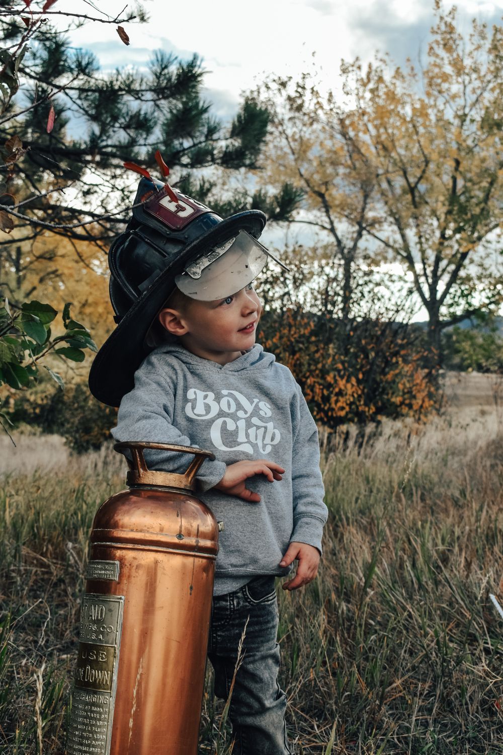 Toddler in gray Boys Club hoodie wearing firefighter helmet stands beside vintage copper fire extinguisher outdoors.