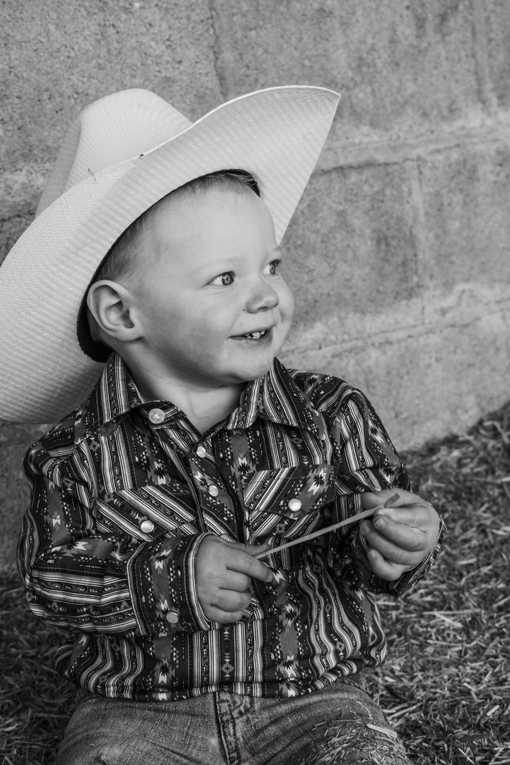 Smiling young boy in cowboy hat and western shirt sitting in hay in black and white photo.