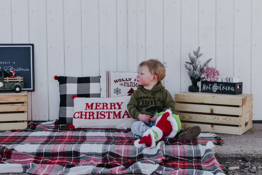 Cute toddler boy sitting on a plaid blanket with Christmas decorations and holiday pillows.