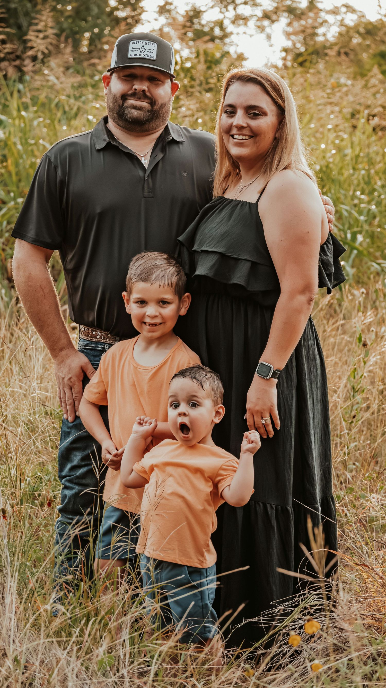 Family of four posing in a golden field, parents in black outfits with two young boys in matching orange shirts.