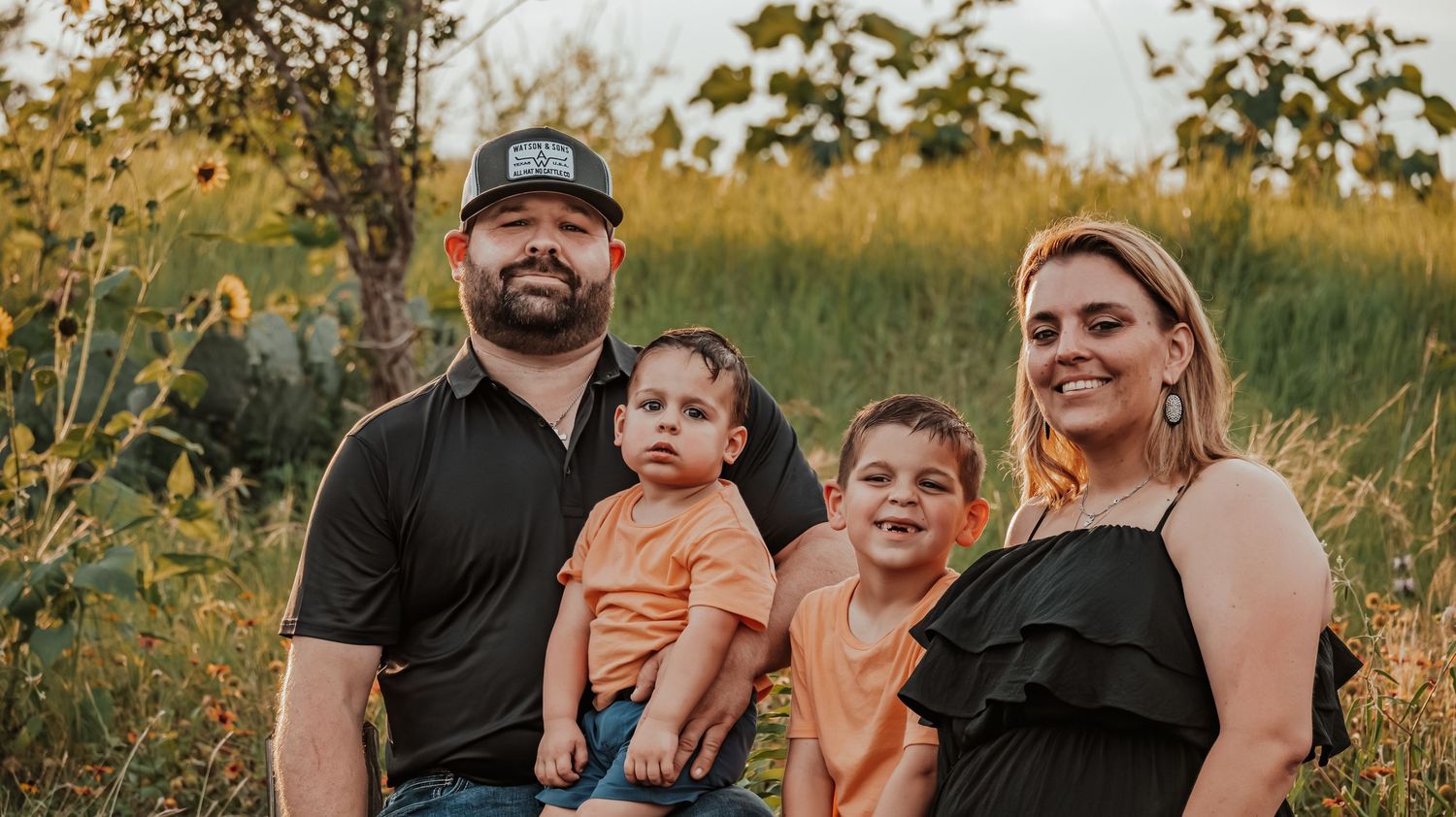A happy family of four poses outdoors in a golden field, with parents holding two young boys in orange shirts.