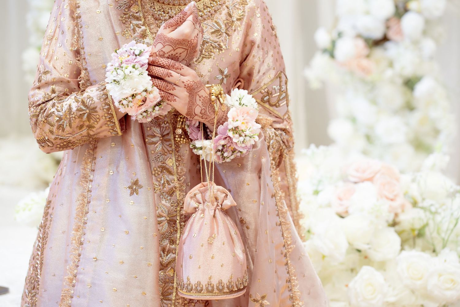 Bride in blush pink embroidered lehenga holding floral accessories at a wedding ceremony.