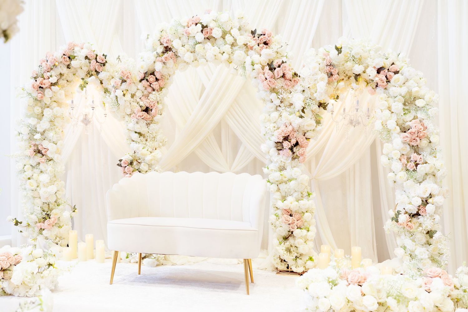 Elegant wedding photo booth with white floral arch, blush roses, draped fabric, and white loveseat with candles.