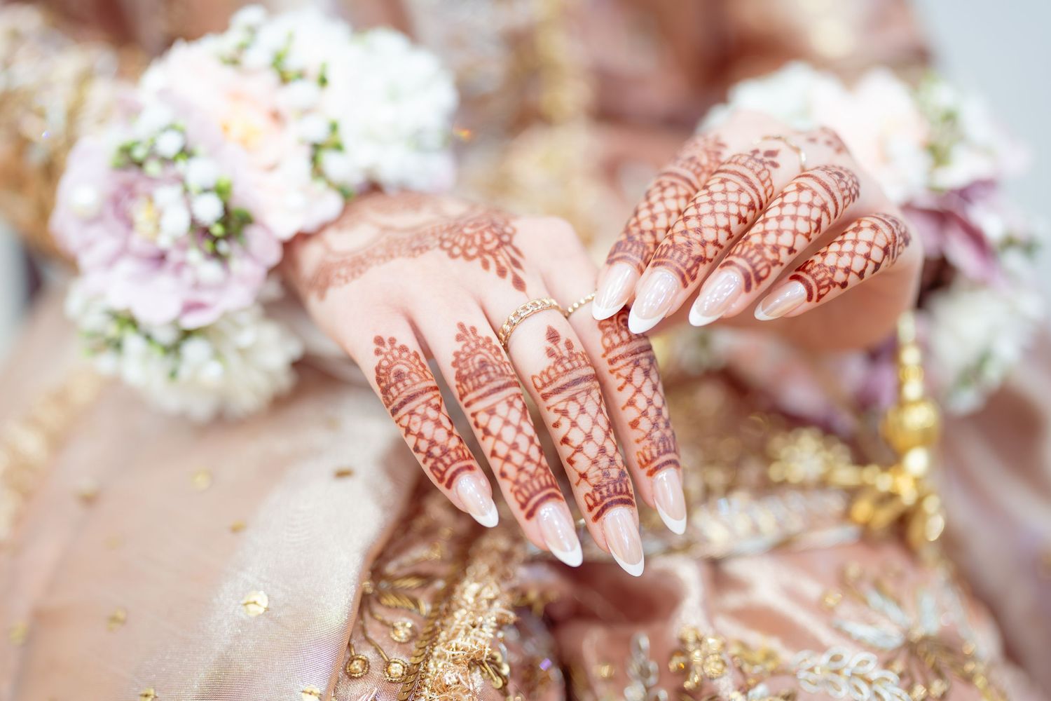 Bride's hands adorned with intricate henna mehndi designs and long white nails, holding a floral bouquet in bridal attire.
