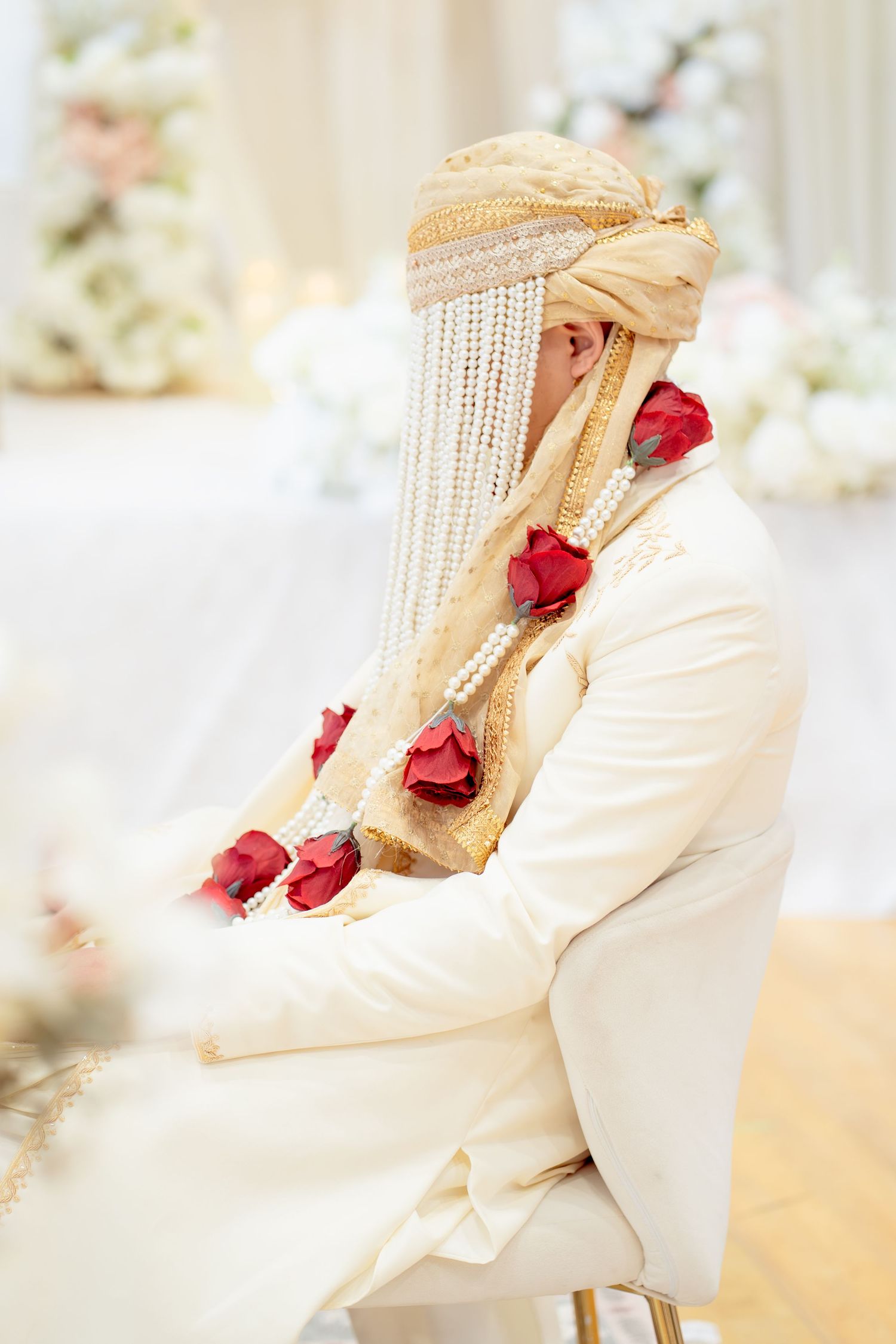 Groom in white suit and gold turban with red roses and pearl garland at Indian wedding ceremony.
