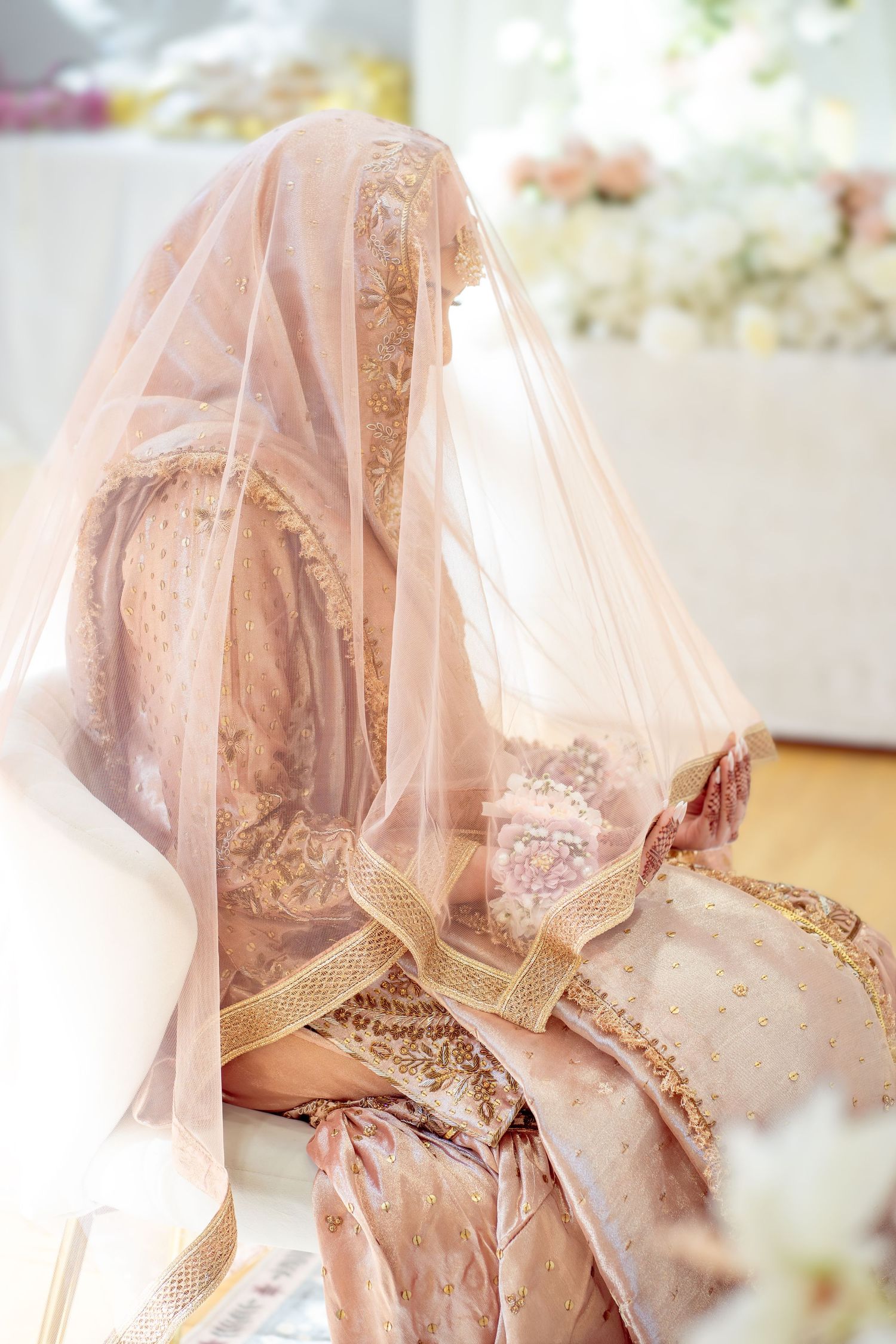 Bride seated in blush pink embroidered Indian bridal lehenga covered with sheer veil at ceremony.