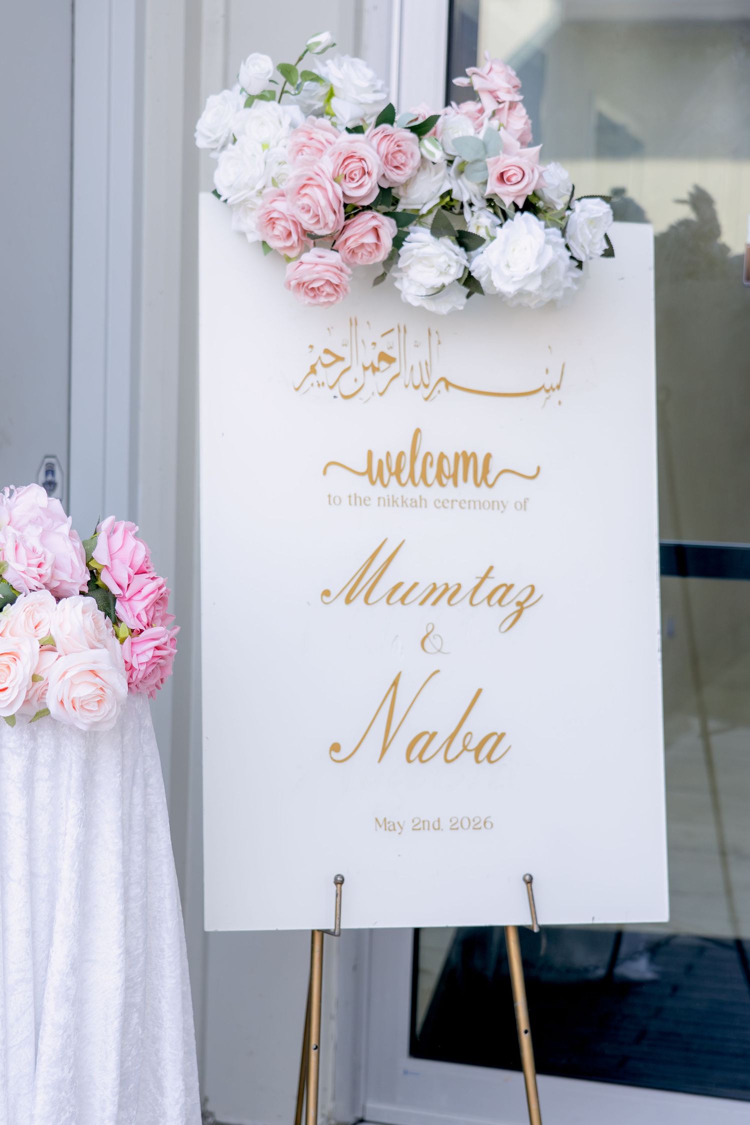 White and gold welcome sign for Mumtaz and Naba Nikkah ceremony adorned with pink and white roses.