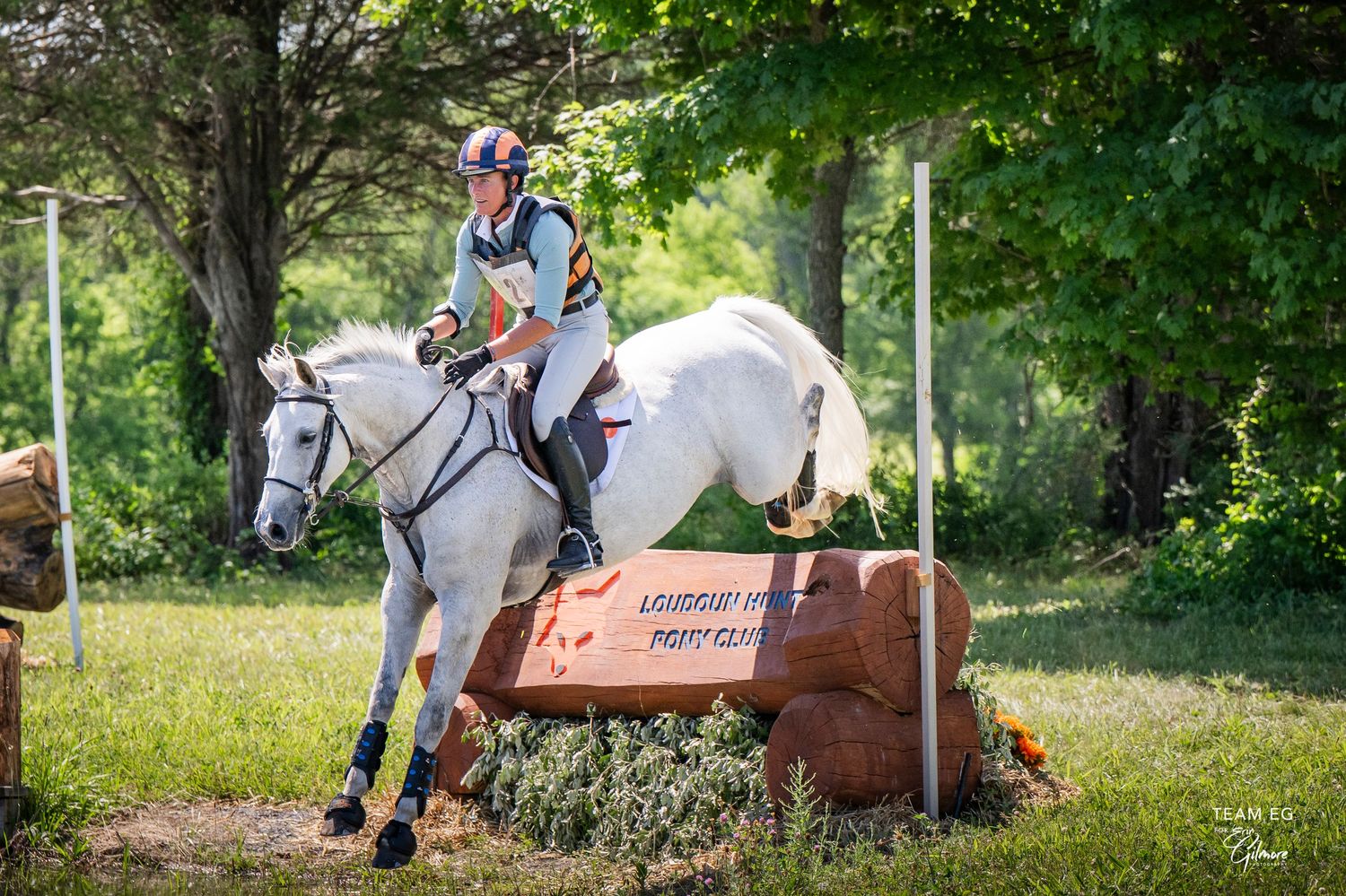 Rider on white grey horse jumping over log obstacle during cross-country equestrian event outdoors.