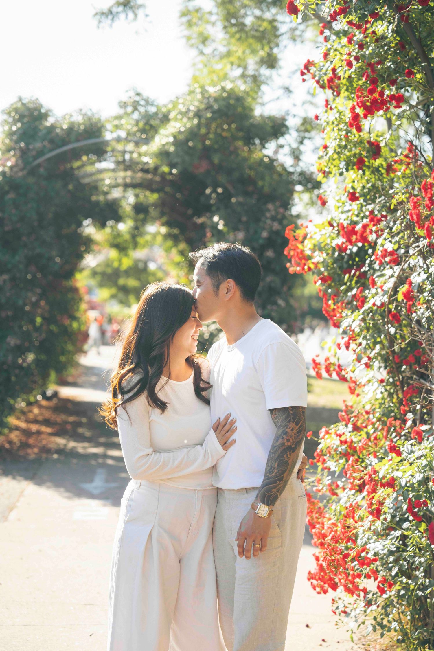 Couple sharing a romantic moment near red bougainvillea flowers in a sunny garden, woman in white outfit, man with tattoos.