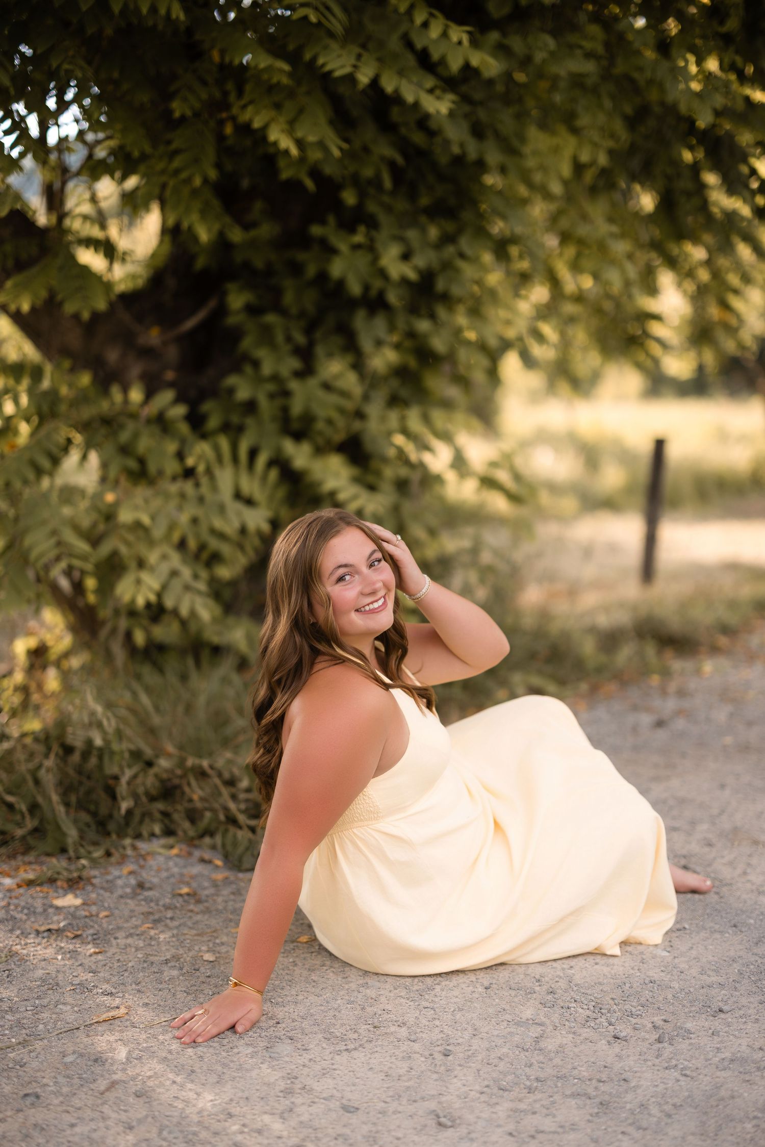 Young woman in yellow dress sitting on ground smiling, surrounded by lush green trees in golden sunlight.