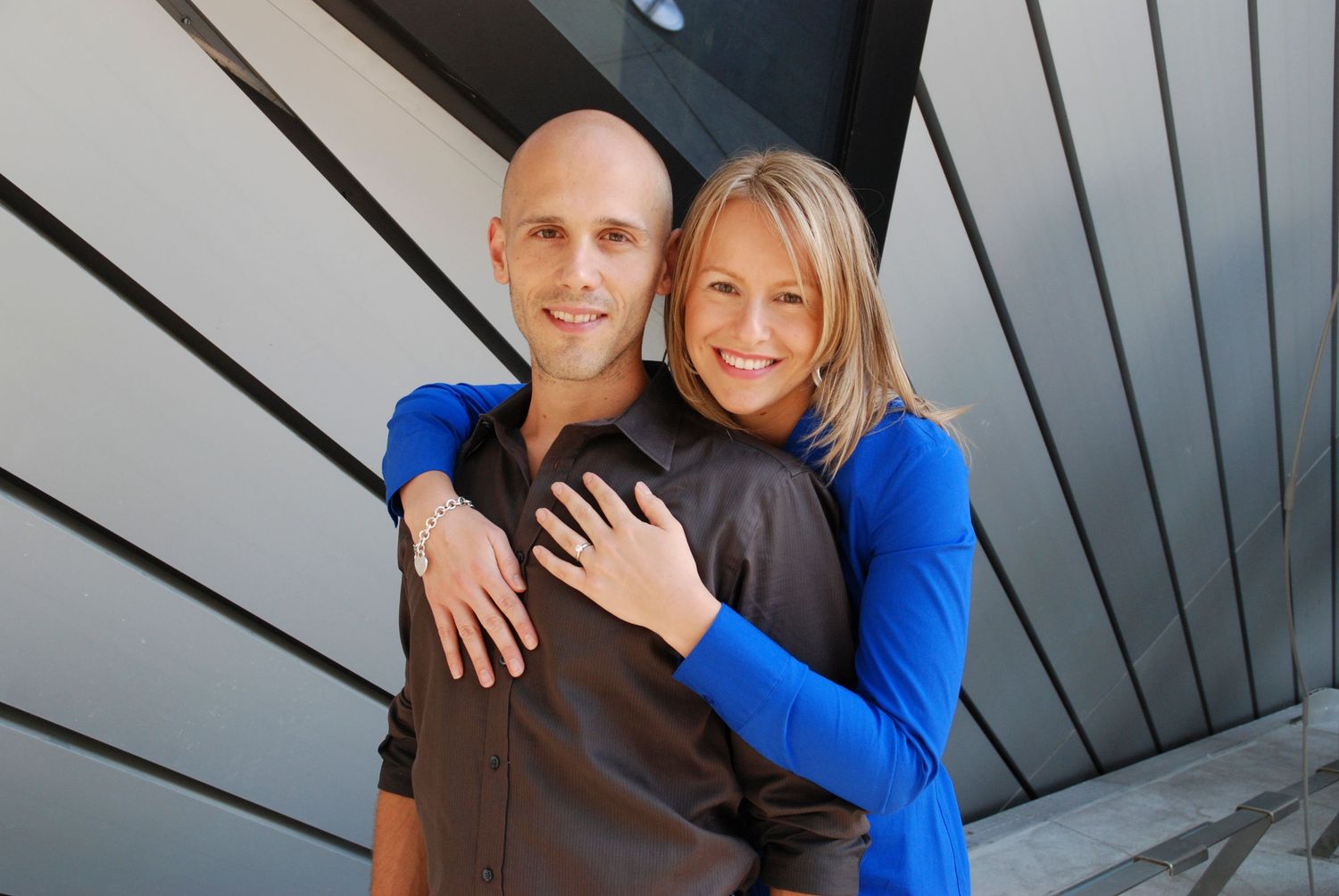 Smiling couple embrace warmly in front of a modern geometric patterned building exterior.