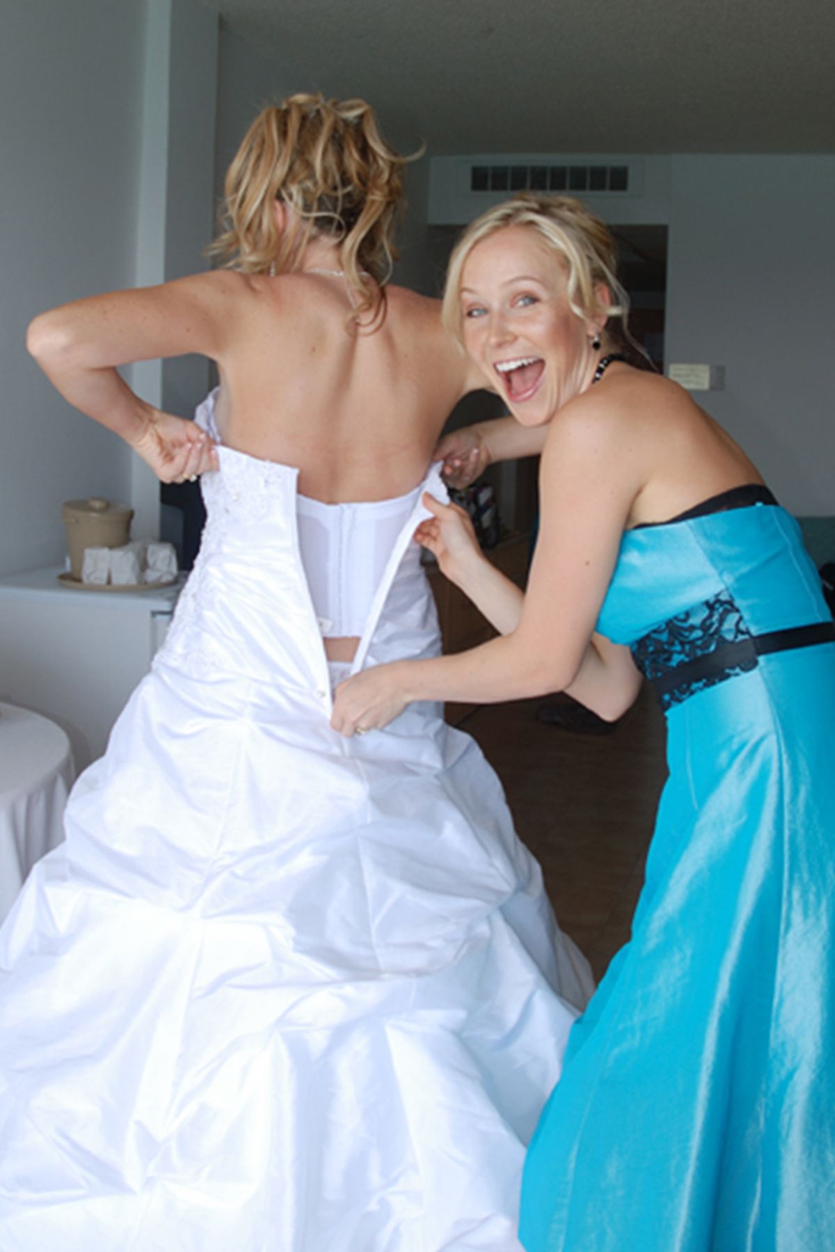 Bridesmaid in turquoise dress excitedly helping bride lace up white wedding gown in hotel room.
