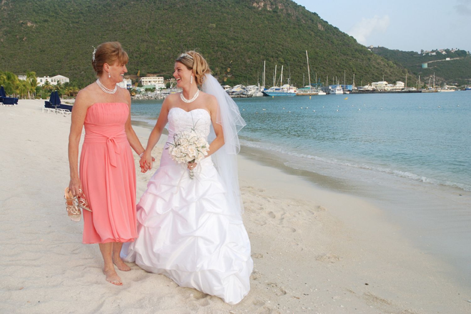 Bride in white strapless gown and bridesmaid in coral dress walking hand in hand on tropical beach wedding.