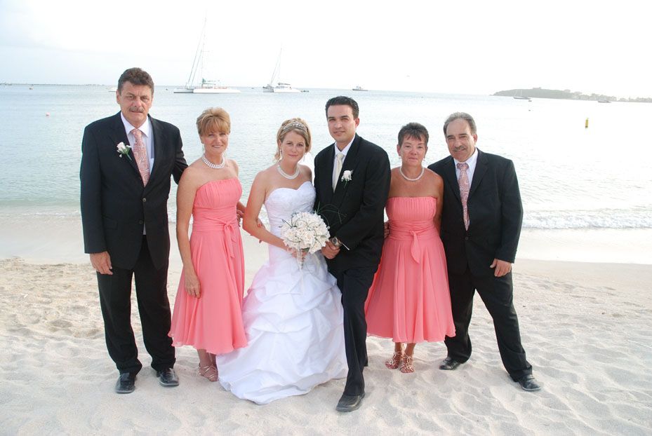 Wedding party posing on a sandy beach, bride in white gown with bridesmaids in coral pink dresses.