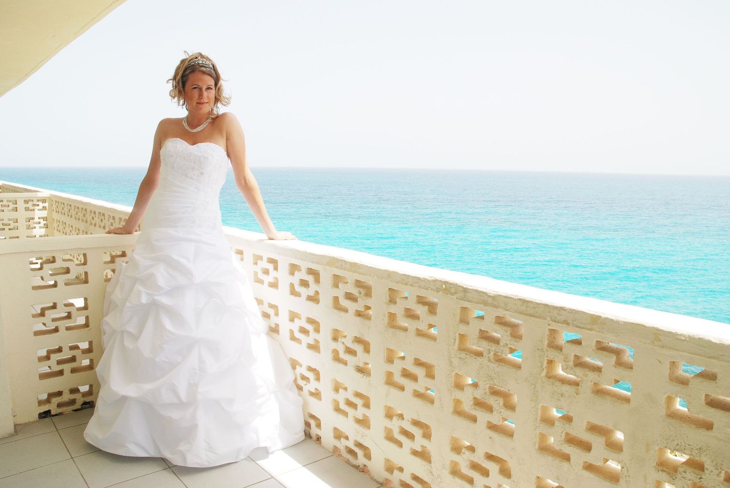 Elegant bride in white ball gown standing on a balcony overlooking the turquoise ocean on her wedding day.