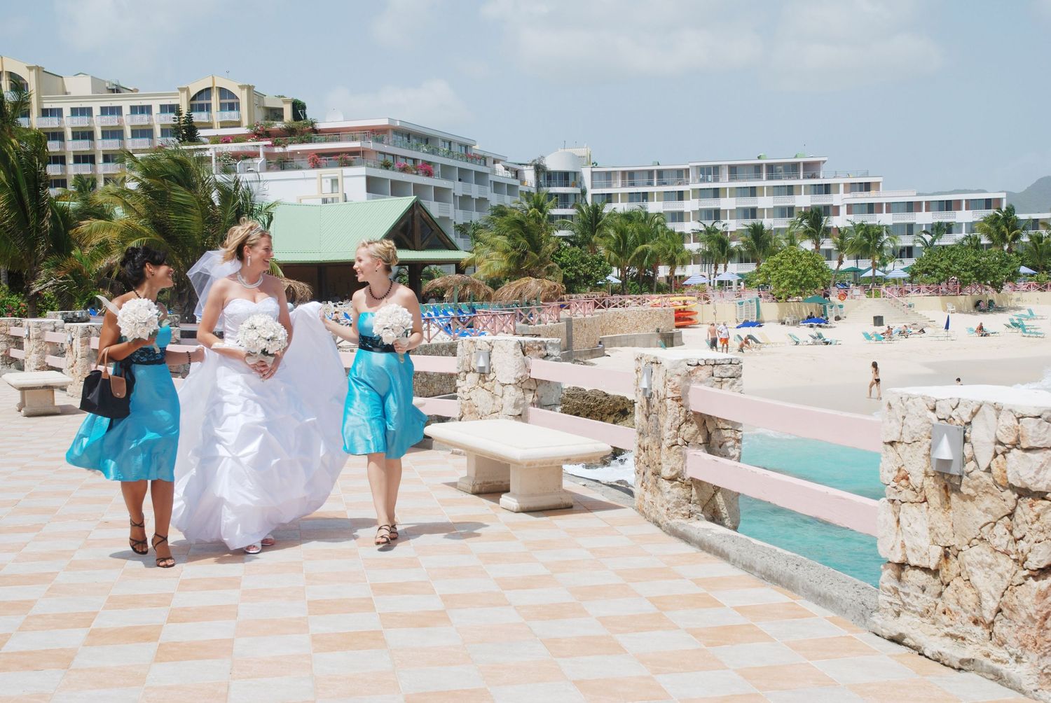 Bride in white gown walks with bridesmaids in turquoise dresses along beachside promenade at resort.