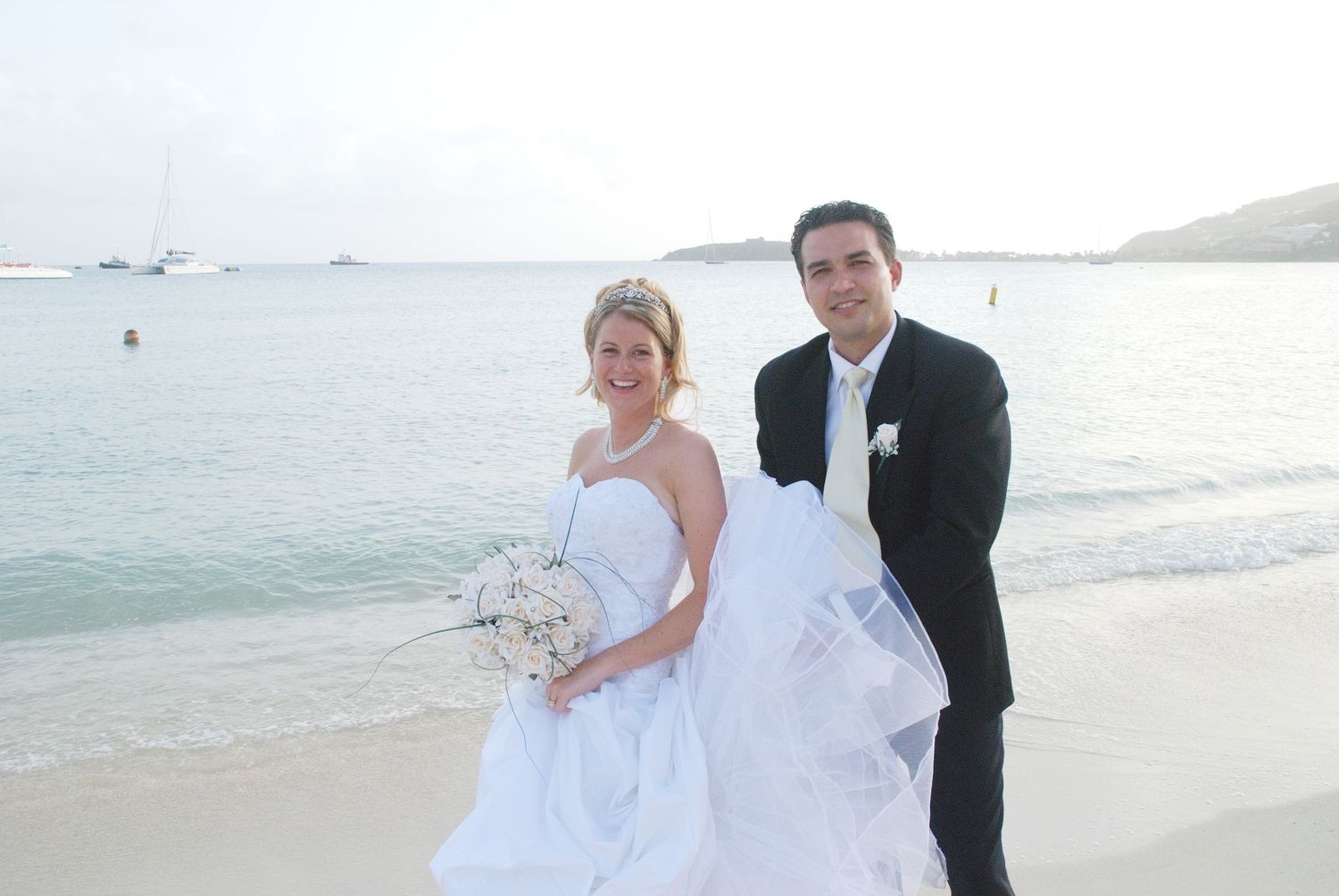 Smiling newlywed couple in wedding attire poses romantically on tropical sandy beach by calm ocean.