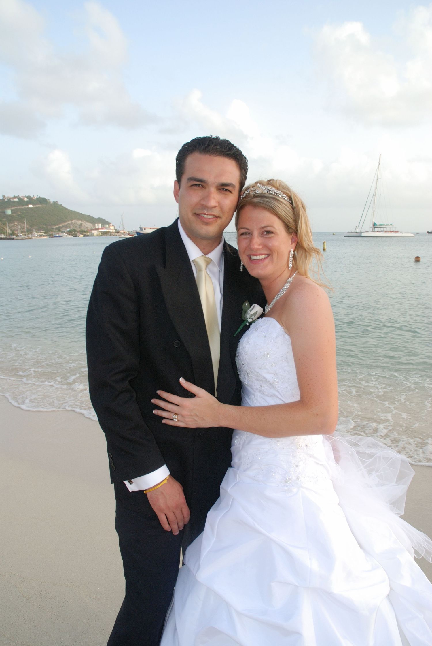 Bride and groom share a romantic moment on a tropical beach with sailboats behind them.
