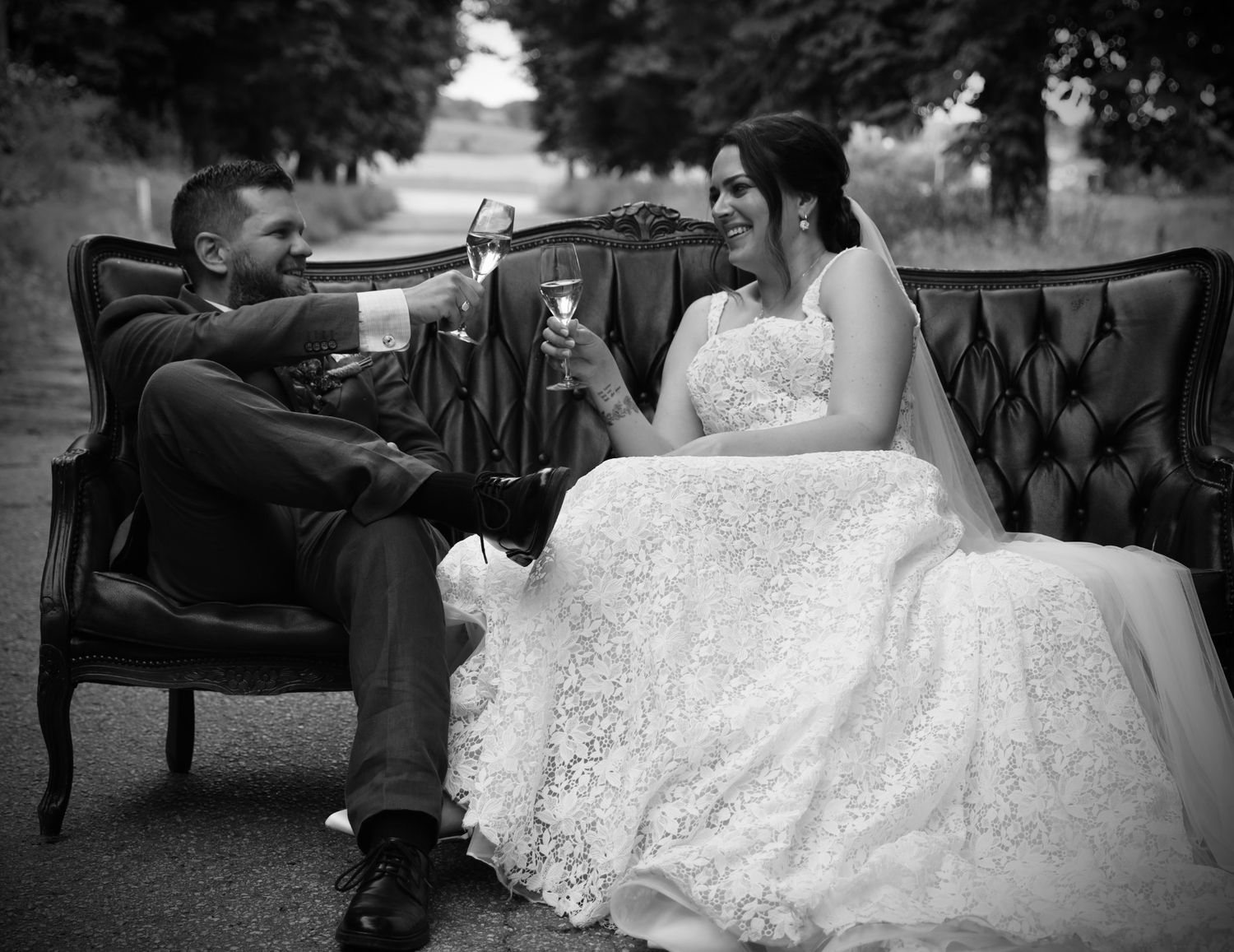 Black and white photo of bride and groom toasting champagne on a vintage leather sofa outdoors.
