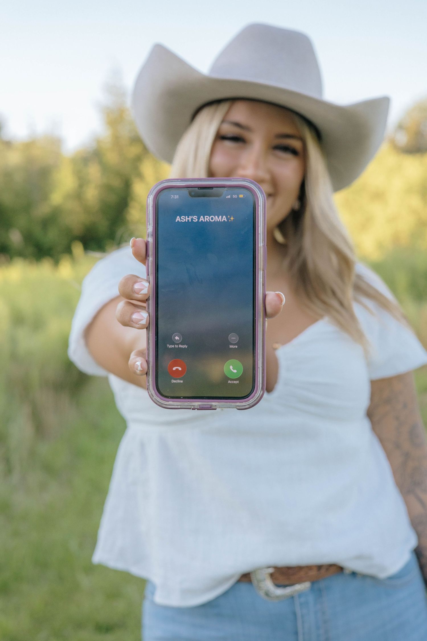 Smiling blonde woman in cowboy hat holding phone showing incoming call from Ashy's Aroma.