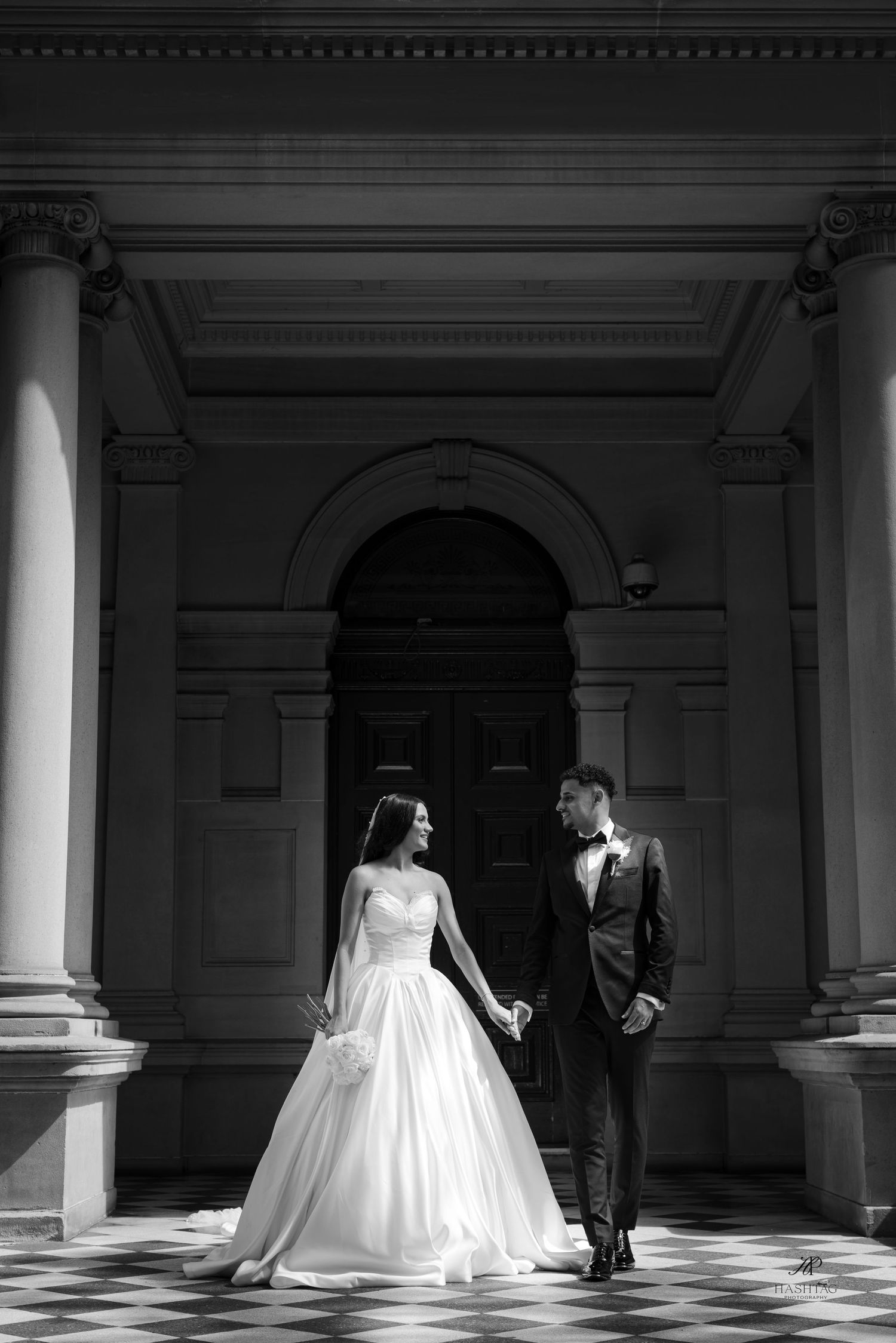 Black and white photo of bride and groom holding hands before grand classical building.