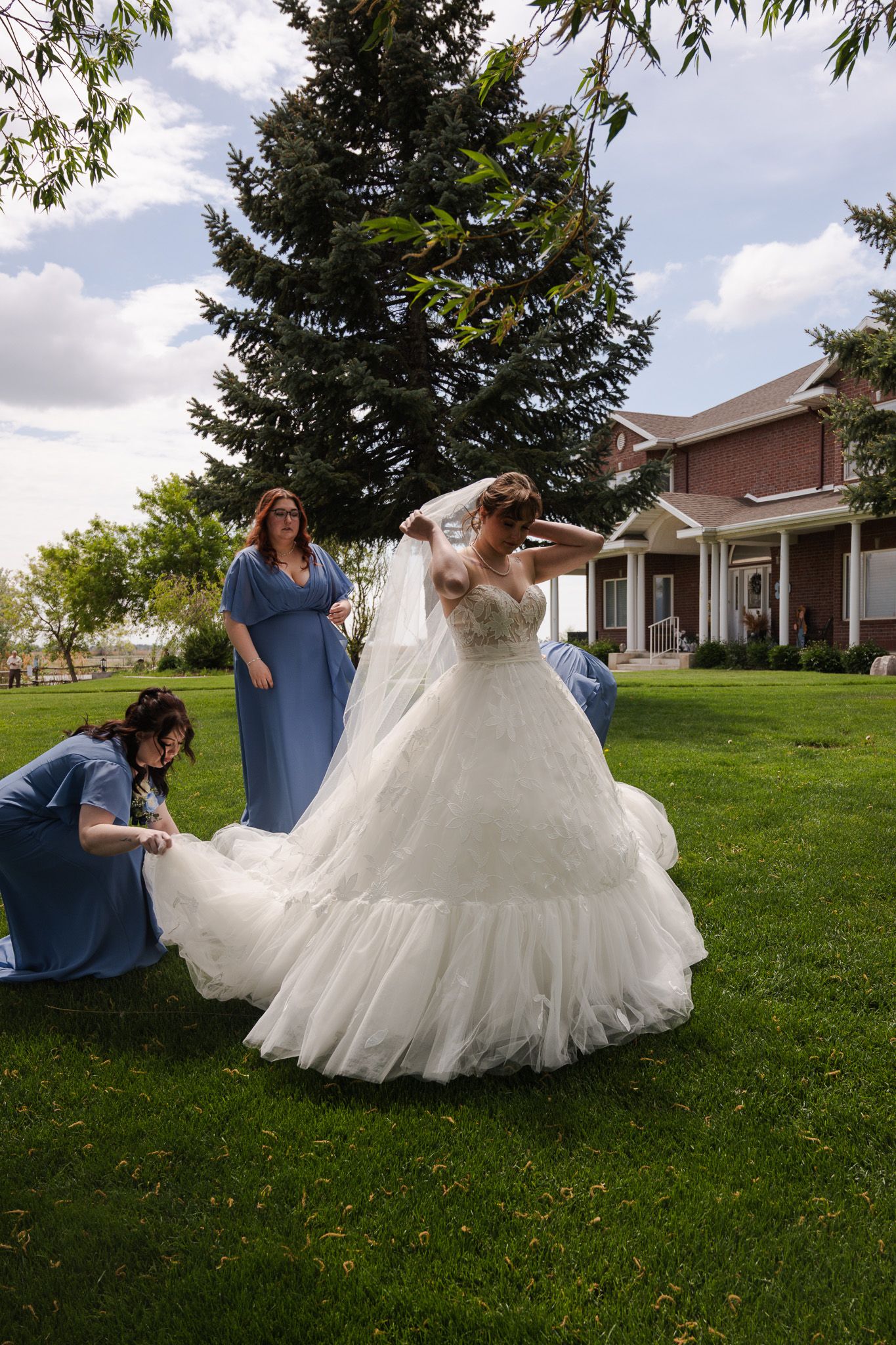 Bride in white ball gown twirls outdoors as bridesmaids in blue dresses adjust her veil and train on green lawn.