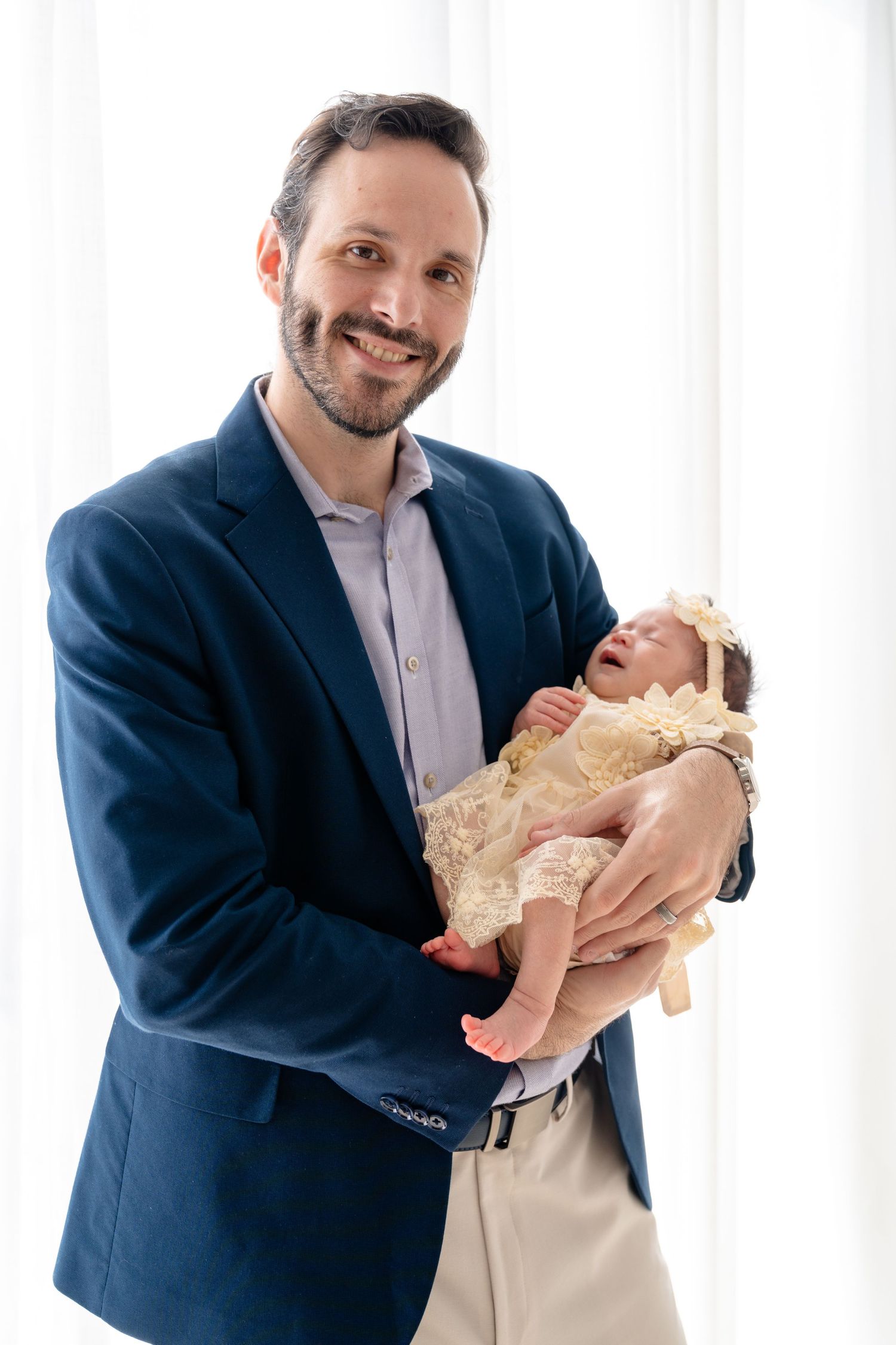 Smiling father in blue blazer holding newborn baby dressed in cream lace outfit with floral headband.
