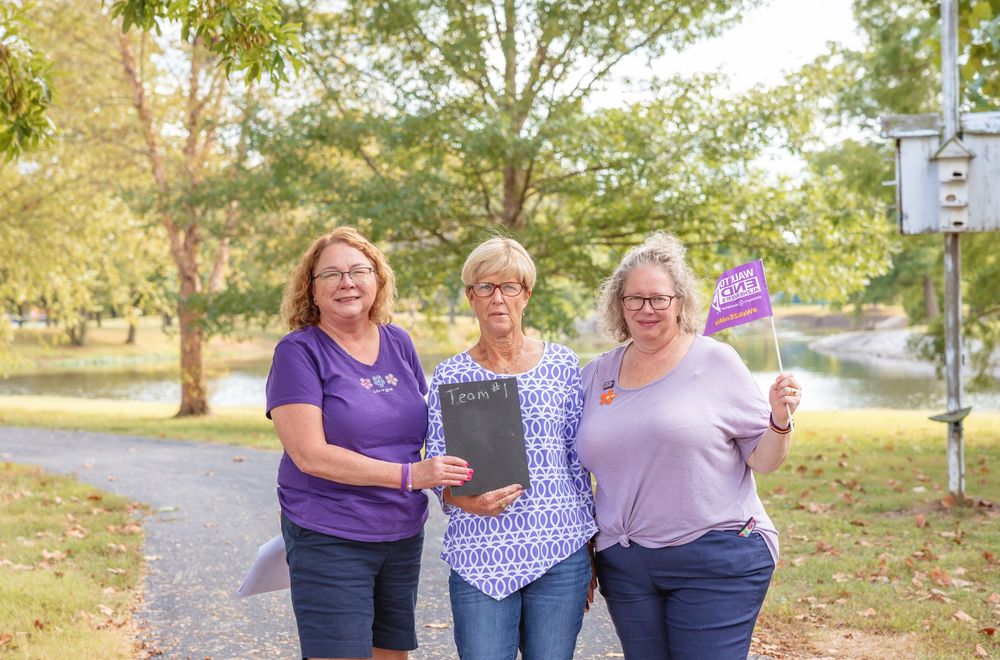 Three women holding purple Alzheimers awareness flags and clipboard on scenic outdoor walking path.