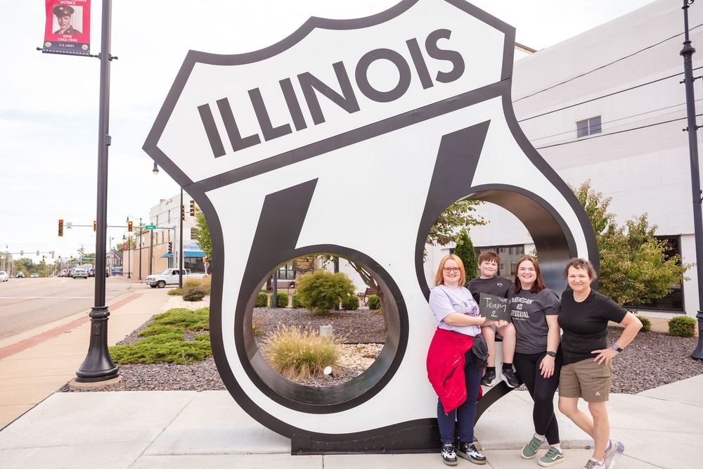 Group of four people posing in front of Illinois Route 66 sign monument on city street.