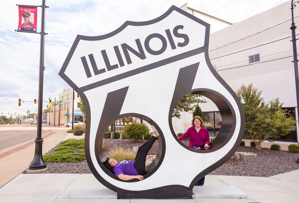 Two people pose inside a large Route 66 Illinois highway sign sculpture on a city sidewalk.