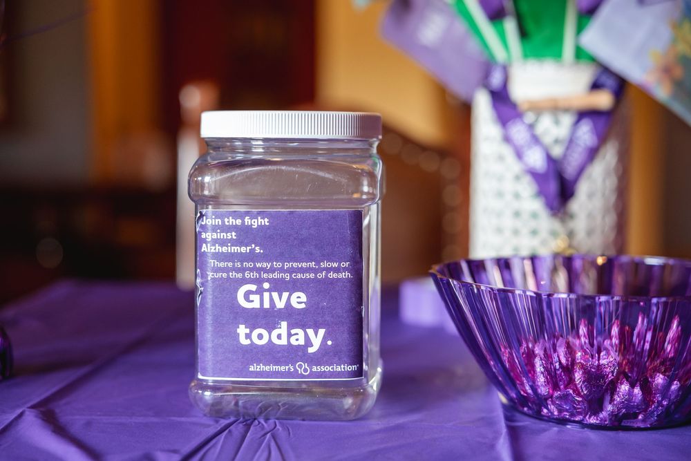 Alzheimer's Association donation jar with purple label saying Give Today on a purple tablecloth.