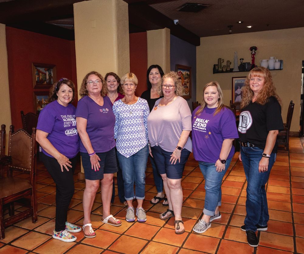 Group of eight women posing together at a charity fundraiser event, several wearing matching purple logo t-shirts.