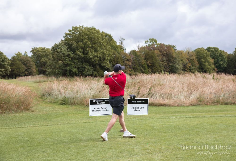 Golfer in red shirt swinging club on lush green golf course with tall grass and sponsor signs in background.