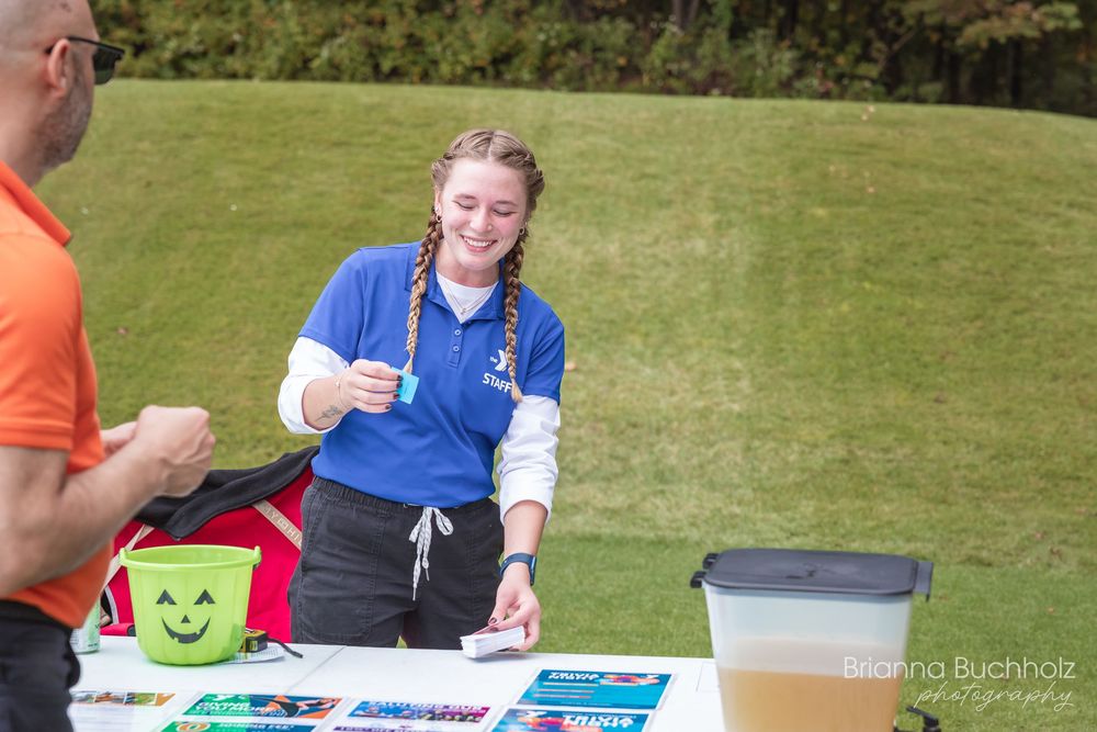 Young woman in blue polo shirt working at outdoor registration table with brochures and supplies on green lawn.