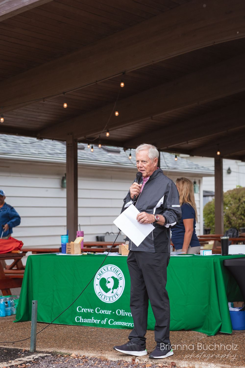 Man speaking at podium with Creve Coeur-Olivette Chamber of Commerce green tablecloth at outdoor event.
