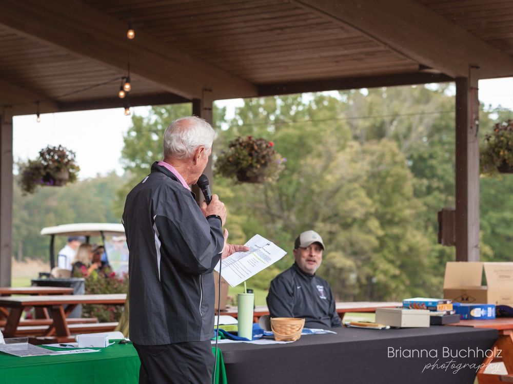 Man speaking at outdoor golf club event podium while attendee sits at registration table under pavilion.