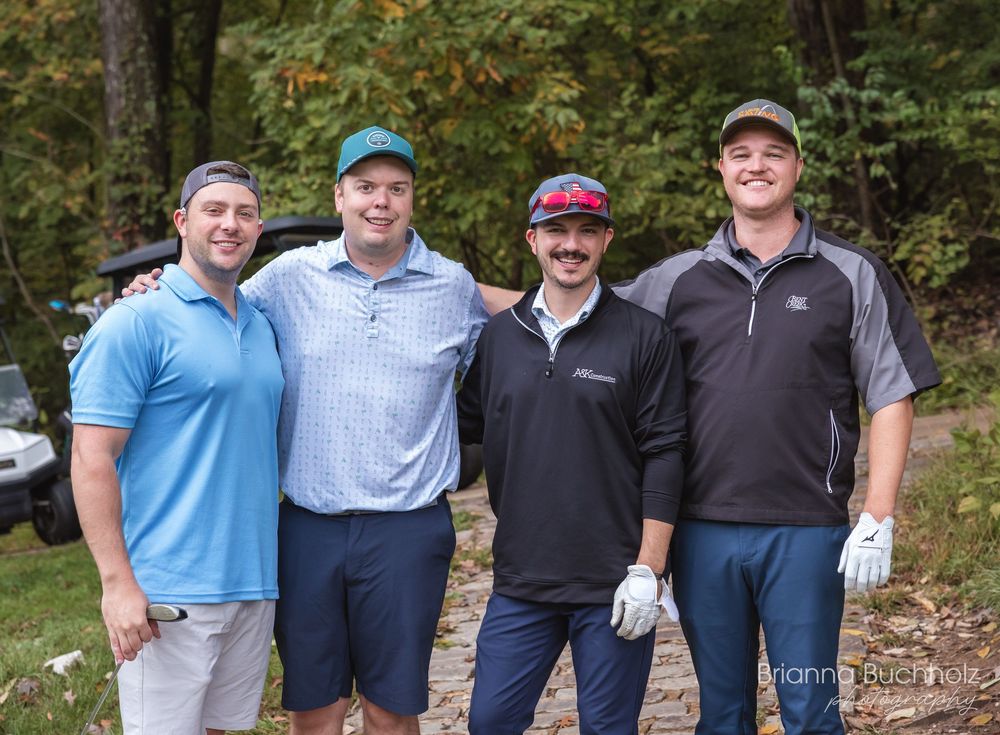 Four men smiling together outdoors at a golf event, surrounded by autumn trees.